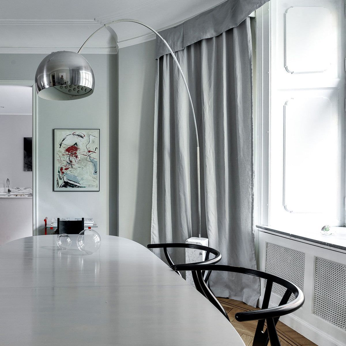 The Clérachic arc lamp's chrome dome hangs low over a pale dining table, its marble base framed by soft grey curtains.
