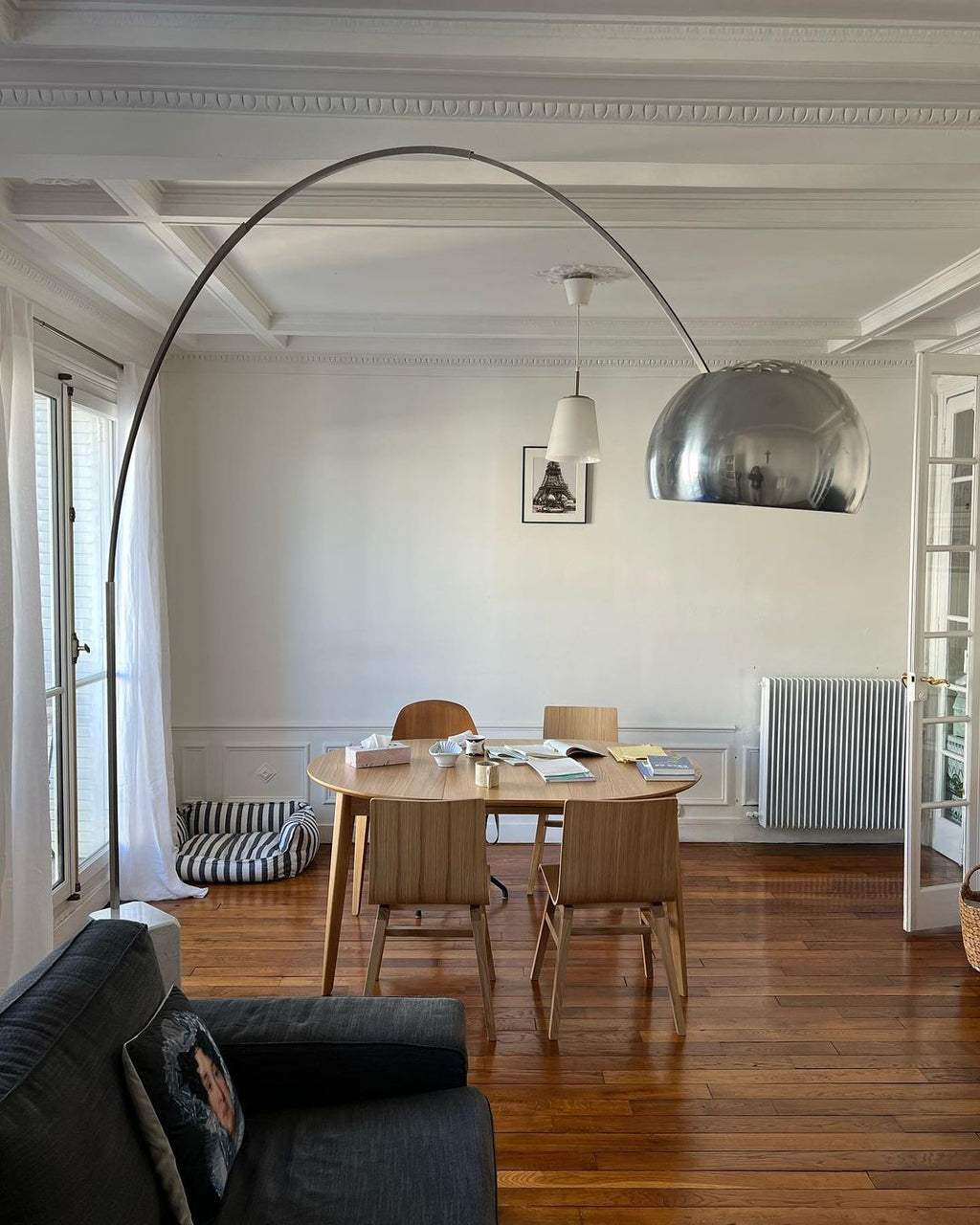 A Clérachic arc floor lamp's chrome dome glows above a wooden dining table set beneath coffered ceilings and dark parquet flooring.