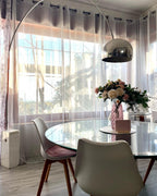 Sunlight streams through sheer curtains as the Clérachic arc lamp's chrome shade hovers over a glass dining table and pink roses.