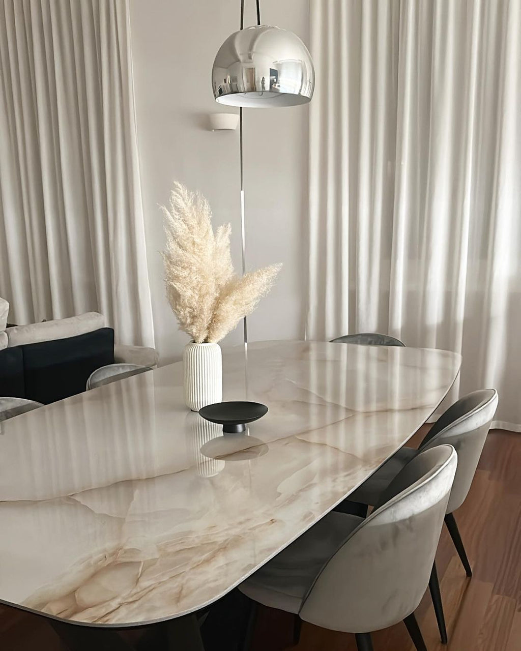 The Clérachic arc lamp's dome hangs above a marble-topped dining table dressed with a pampas grass arrangement and grey chairs.