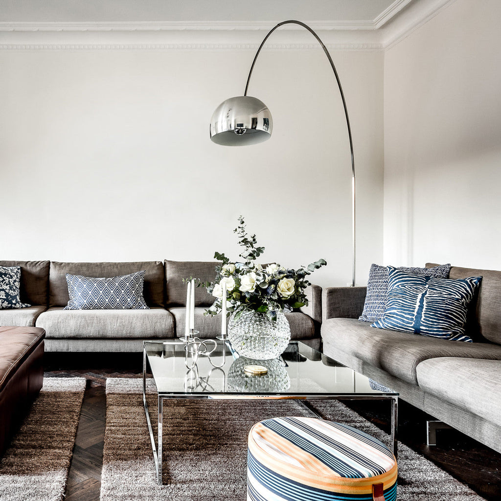 Clérachic's arcing floor lamp reaches over a grey corner sofa and glass coffee table, its chrome dome centred above fresh white roses.