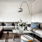 Clérachic's arcing floor lamp reaches over a grey corner sofa and glass coffee table, its chrome dome centred above fresh white roses.