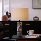 A cup set on a book and a carved wooden sculpture sit beside the Clérachic marble lamp on a dark sideboard.