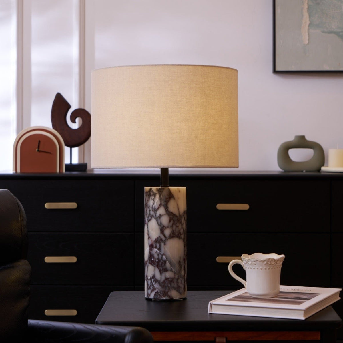 A cup set on a book and a carved wooden sculpture sit beside the Clérachic marble lamp on a dark sideboard.