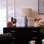 Beside a black leather chair, the Clérachic marble lamp lights a dark sideboard with flowers and a wooden sculpture nearby.