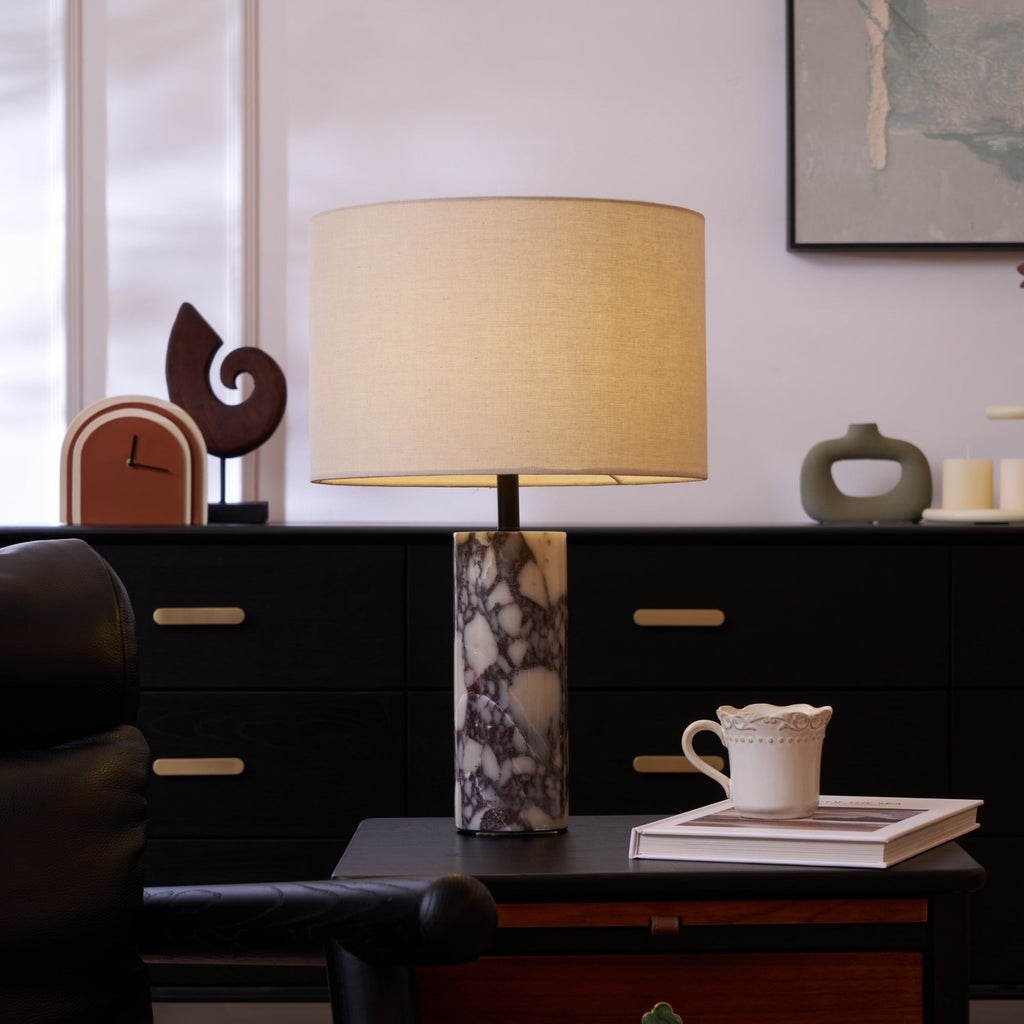 Clérachic marble table lamp on a dark sideboard beside a black leather chair arm, a ruffled cup set on an open book.