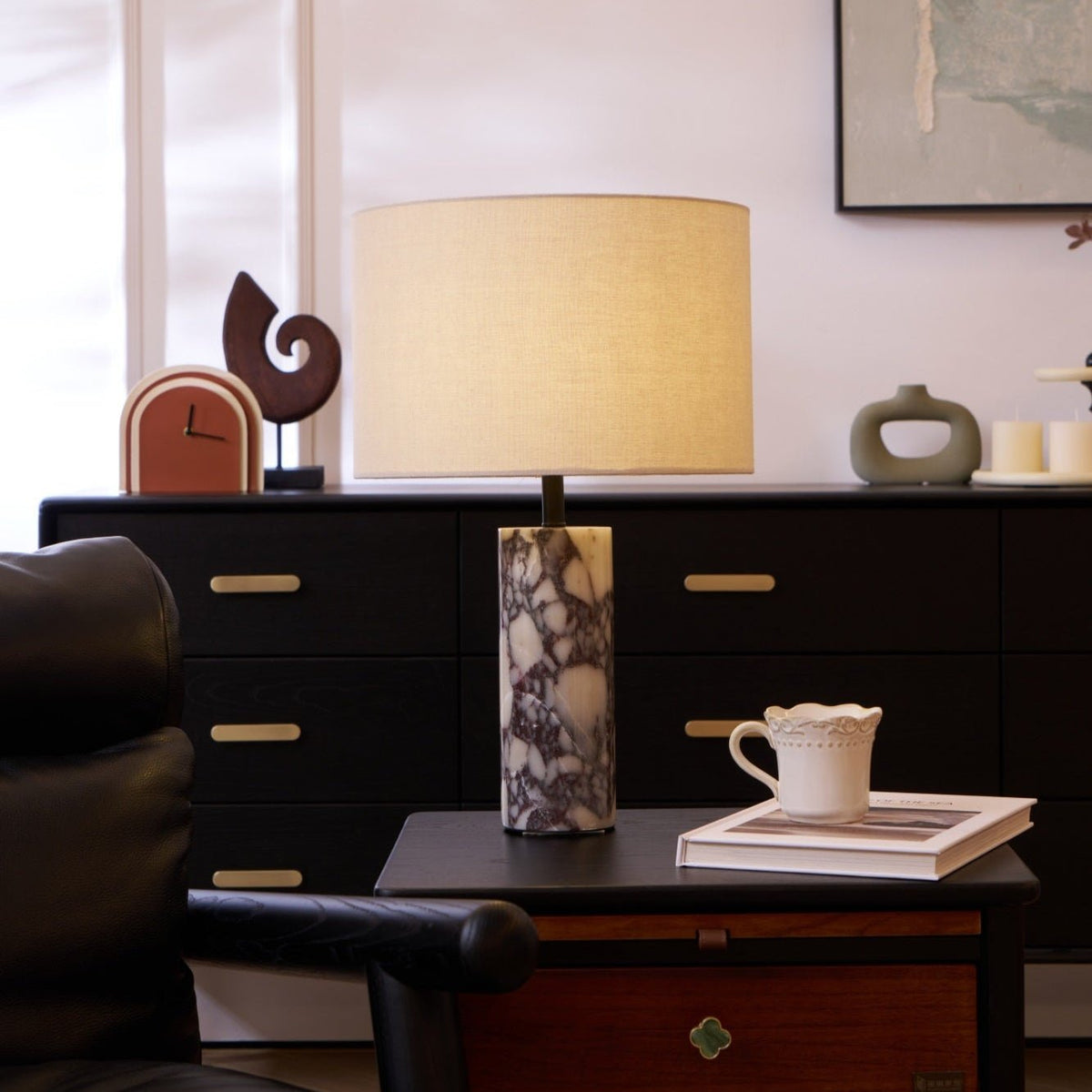 Clérachic marble table lamp on a dark sideboard beside a black leather chair, a cup on a book and a small clock ornament.