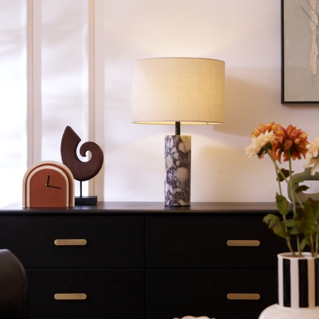 A striped vase of orange and white flowers stands with a clock ornament beside the Clérachic marble lamp on its sideboard.