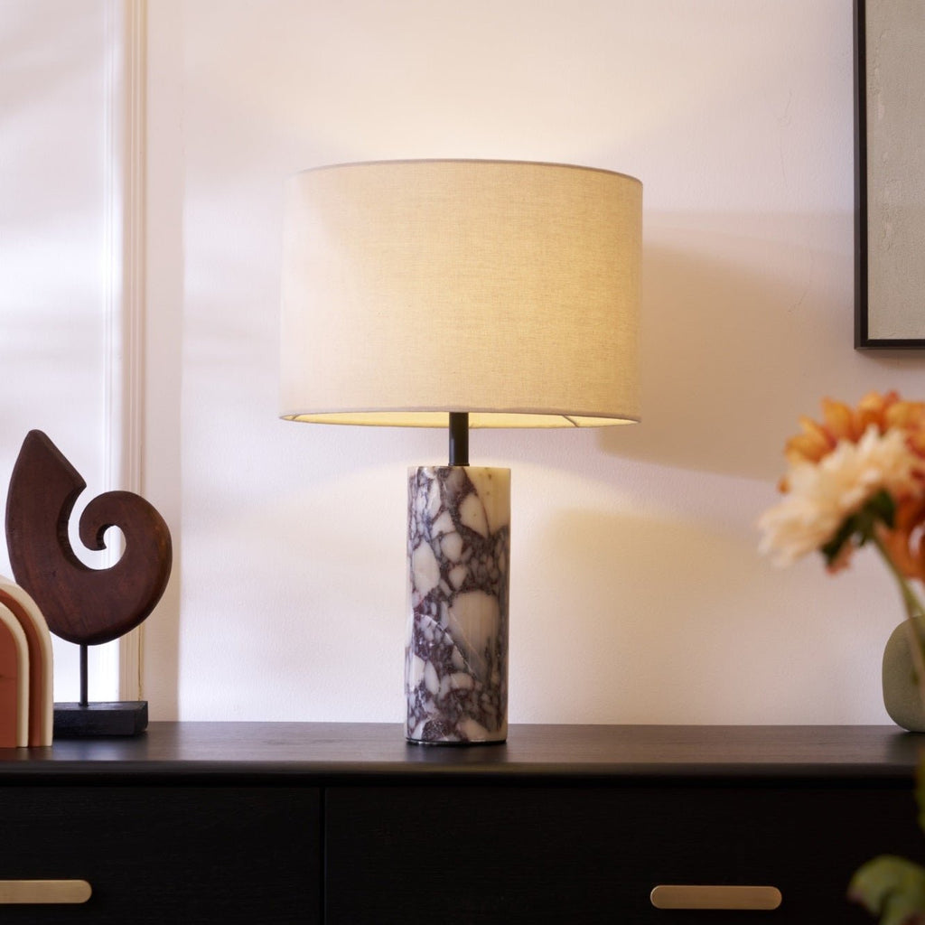 Clérachic marble table lamp lit on a dark wood sideboard, with orange flowers and a carved wooden sculpture nearby.