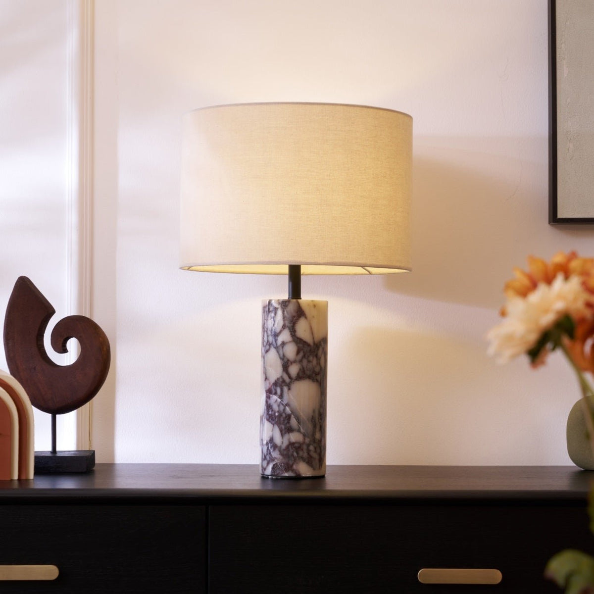Clérachic marble table lamp lit on a dark wood sideboard, with orange flowers and a carved wooden sculpture nearby.
