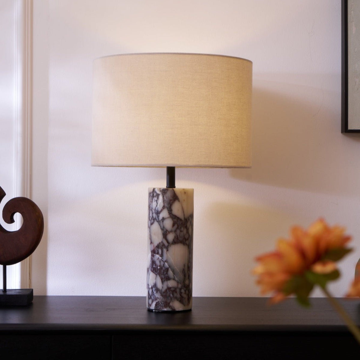 Clérachic marble table lamp lit on a dark sideboard, an orange flower blurred close to the camera on the right.