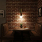 Clérachic wall sconce lit above a leather booth table against an exposed brick wall, with a small plant on the table.