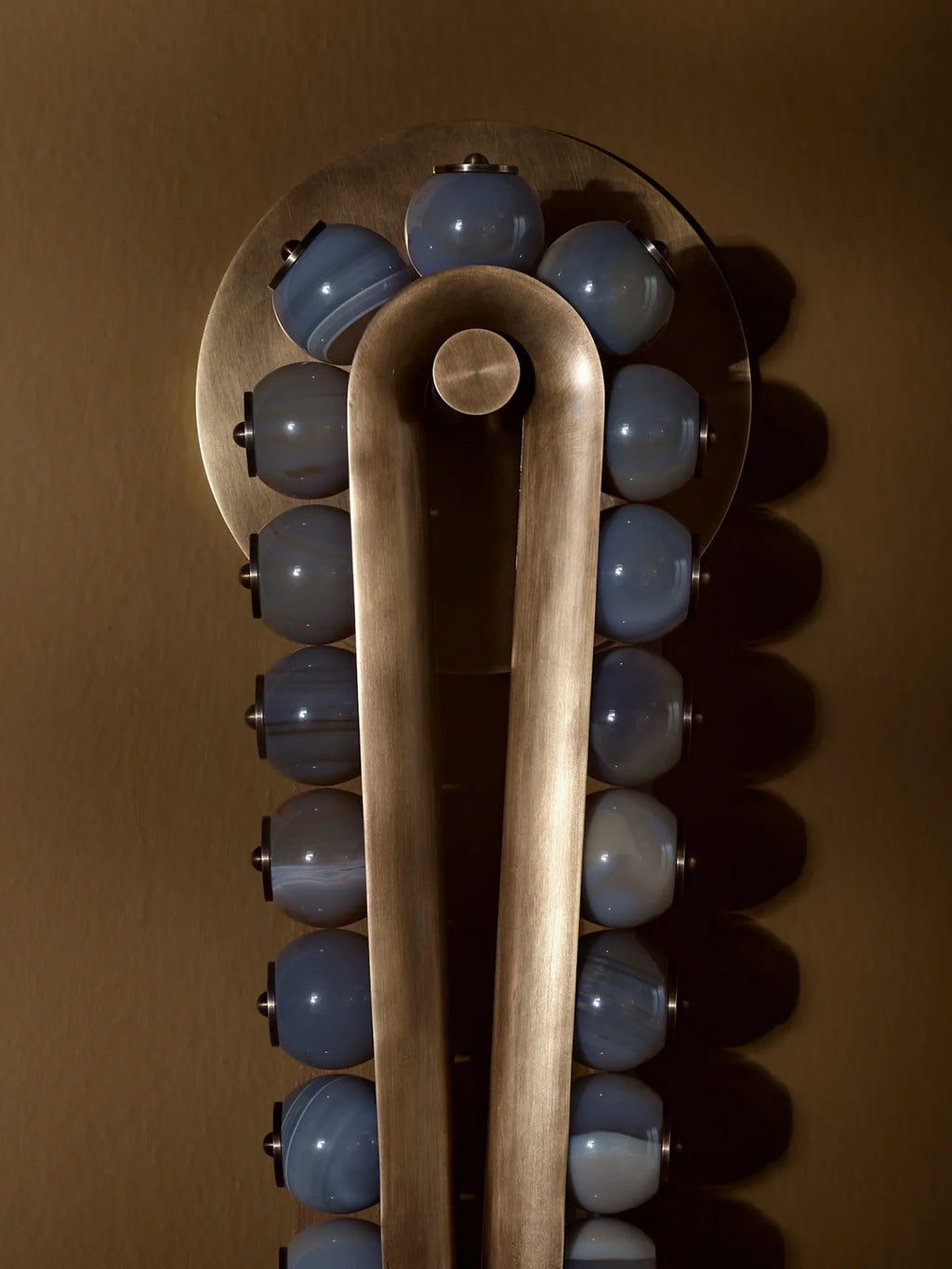 Blue-grey stone beads catch the light along the brass frame of a Clérachic wall sconce, set against a bronze backdrop.