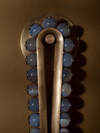 Blue-grey stone beads catch the light along the brass frame of a Clérachic wall sconce, set against a bronze backdrop.