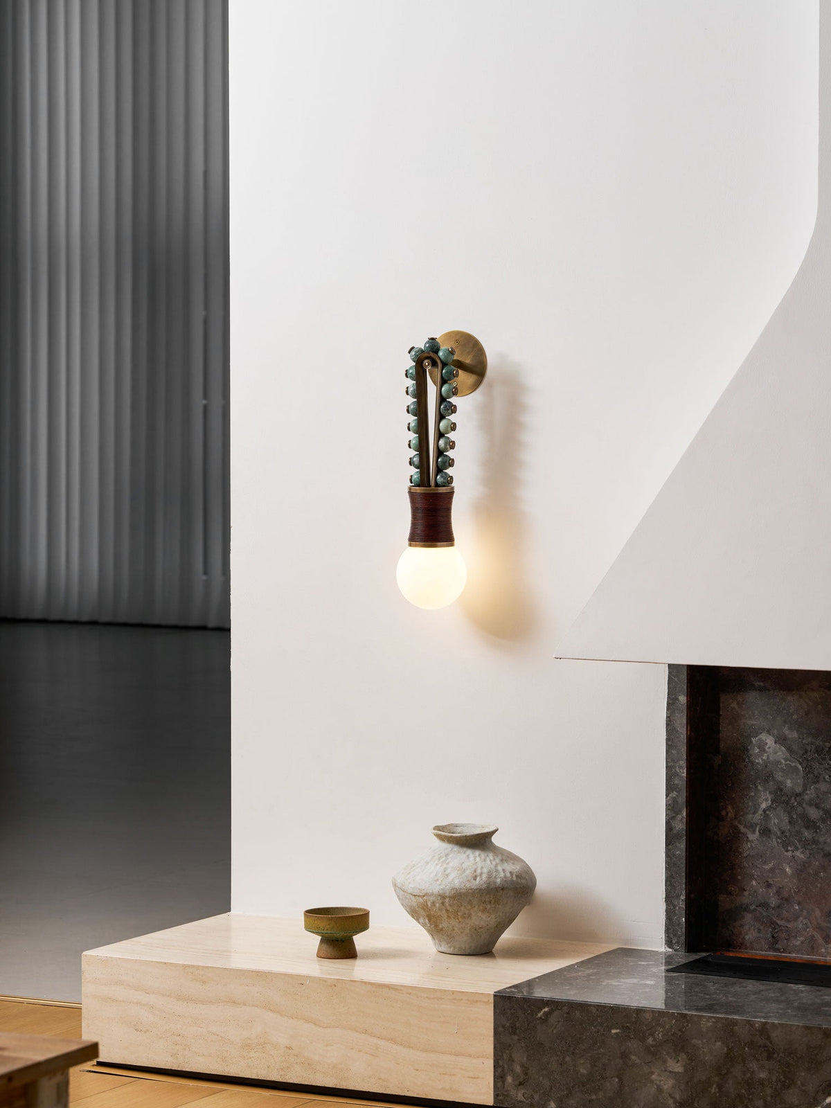 Below a fireplace hearth, a ceramic vase rests on the stone ledge lit by a Clérachic wall sconce in the corner.