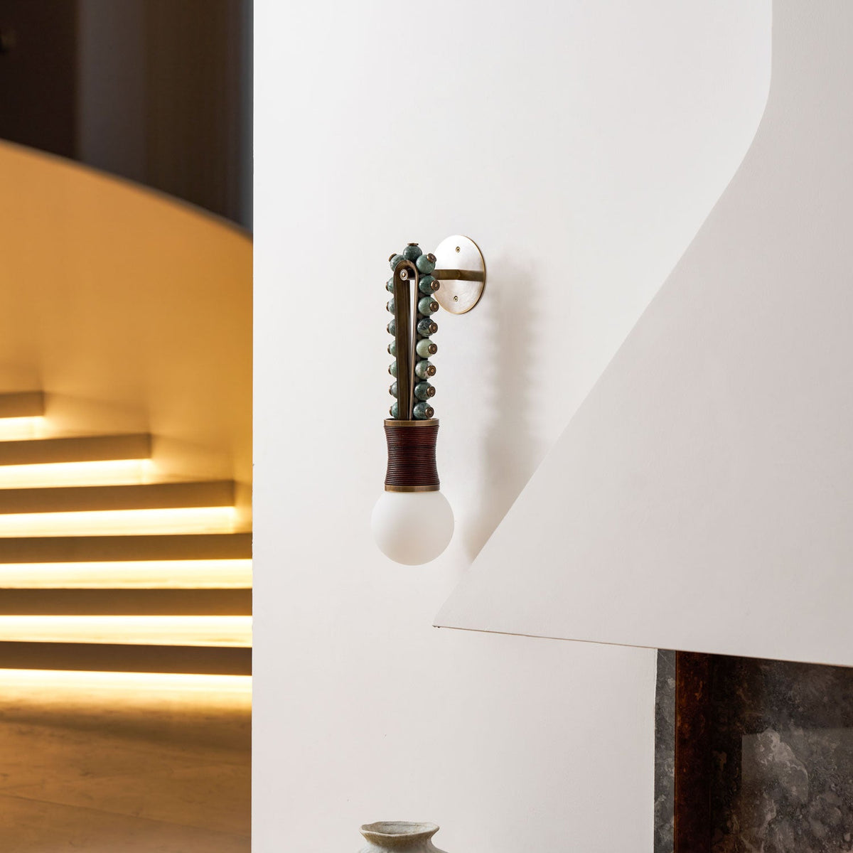 Clérachic wall sconce mounted beside a curved staircase, its pale globe unlit against the white wall and stairwell glow.