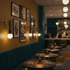 A gallery of framed prints lines a mustard-yellow wall in a restaurant booth, lit by a Clérachic wall sconce.