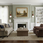 A framed landscape painting hangs above a white mantel, flanked by two lit Clérachic wall sconces in a sage-green room.
