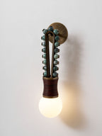 Clérachic wall sconce lit straight-on against a plain white wall, sage-green beads over a maroon-wrapped base.