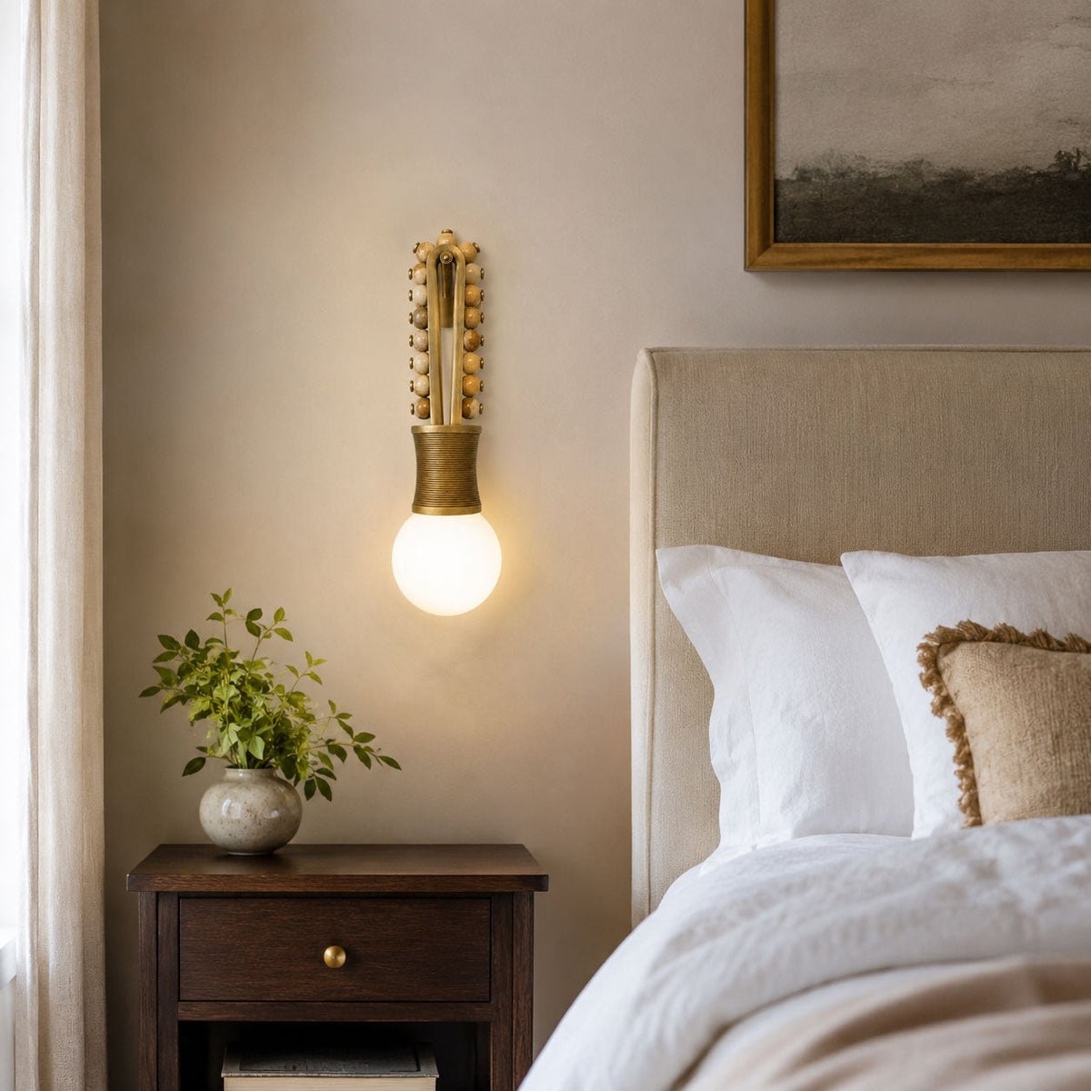 Clérachic wall sconce lit above a bedside table with a small plant, beside an upholstered bed headboard.