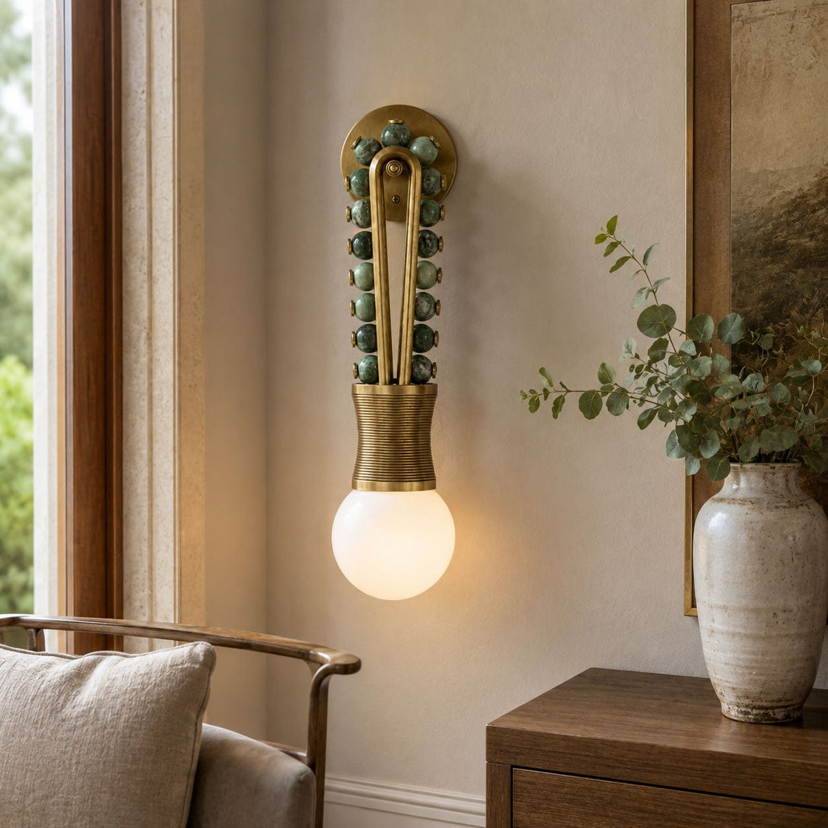 Clérachic wall sconce lit beside a window, olive-toned beads and a brass-wrapped base near a potted plant and armchair.