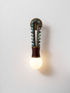 Clérachic wall sconce lit on a plain white wall, sage-green beads and a red-brown wrapped base glowing softly.