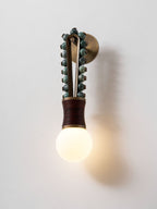 Clérachic wall sconce lit on a plain wall, its sage-green beads and maroon-wrapped base casting a soft shadow beside the glowing bulb.