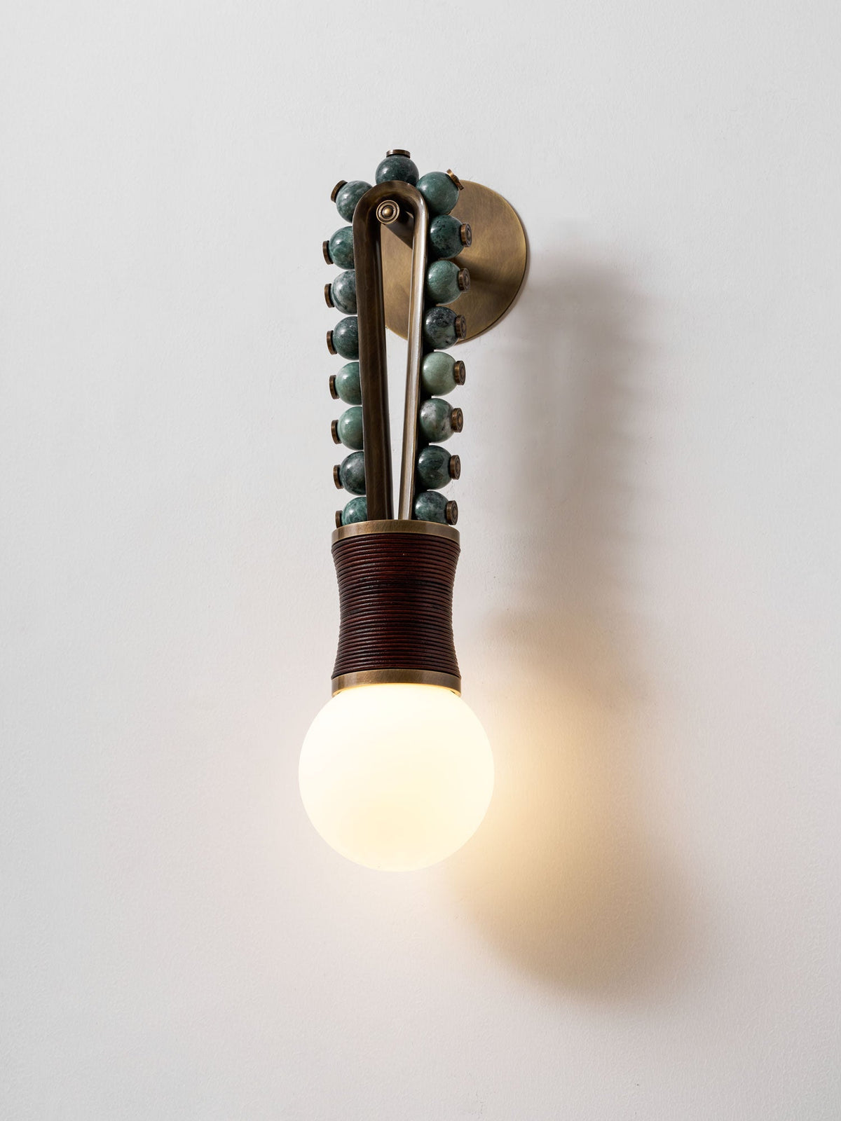 Clérachic wall sconce lit on a plain wall, its sage-green beads and maroon-wrapped base casting a soft shadow beside the glowing bulb.