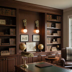 Two Clérachic wall sconces lit either side of a bookshelf nook, glowing above a wood desk in a panelled study.