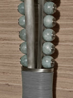Close-up of a Clérachic wall sconce's pale sage stone beads on a metal rod with brass end caps, against a wood-grain wall.