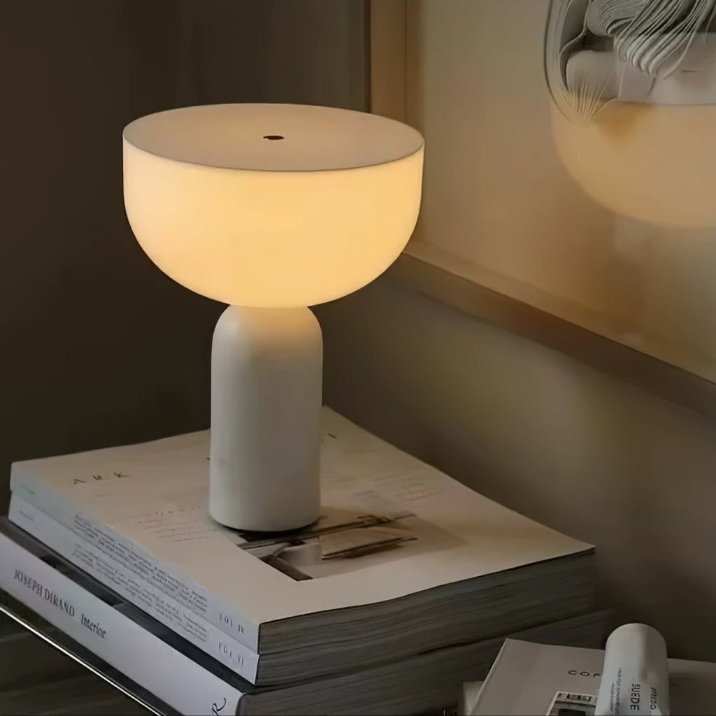 A white marble Clérachic lamp glows warmly on a stack of books beside a window in a dimly lit room.
