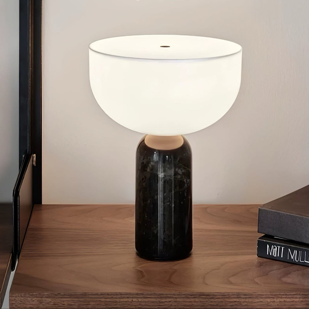 A black marble Clérachic table lamp lit on a walnut console shelf beside a stack of books near an open door.