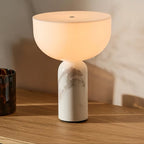 Clérachic's white marble sphere lamp lit on a sunlit wood table beside a dark glass vase and warm daylight.