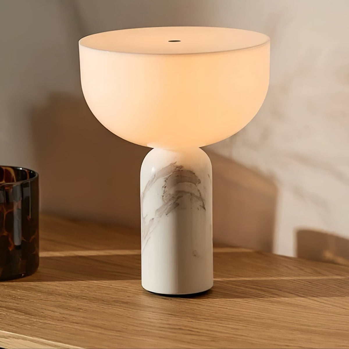 Clérachic's white marble sphere lamp lit on a sunlit wood table beside a dark glass vase and warm daylight.