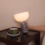 A hand steadies the grey marble base of this Clérachic sphere table lamp beside a teacup and saucer on a dark table.