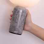 Close-up of a hand gripping the textured grey marble base of this Clérachic table lamp, its dome softly lit above.