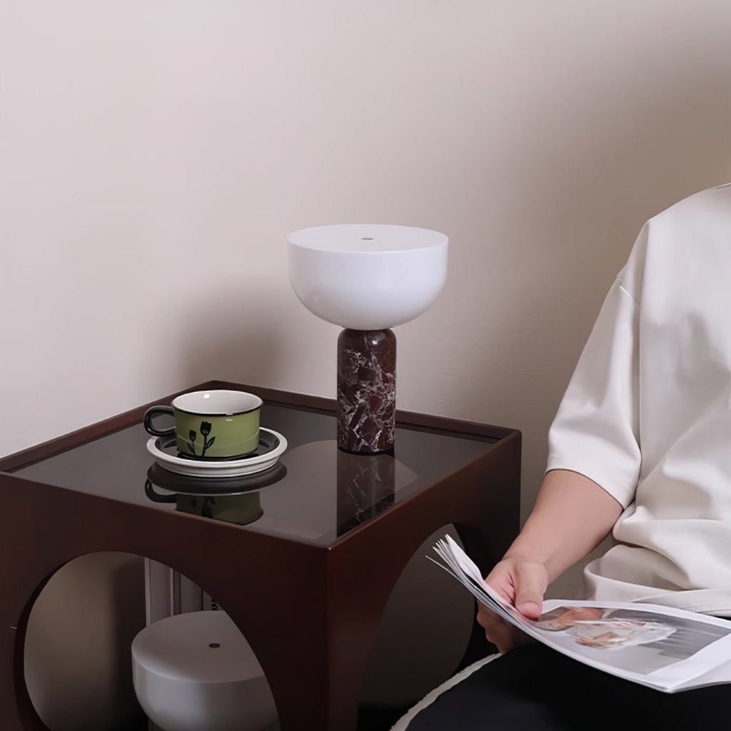 A dark red marble Clérachic lamp lit on a glass side table while someone reads nearby with a cup of tea.