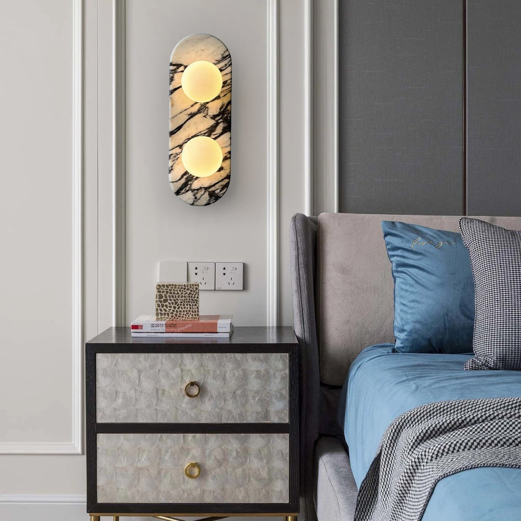 Clérachic marble wall light lit above a dark upholstered nightstand, beside a bed with blue linens and patterned cushions.