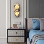 Clérachic marble wall light lit above a dark upholstered nightstand, beside a bed with blue linens and patterned cushions.
