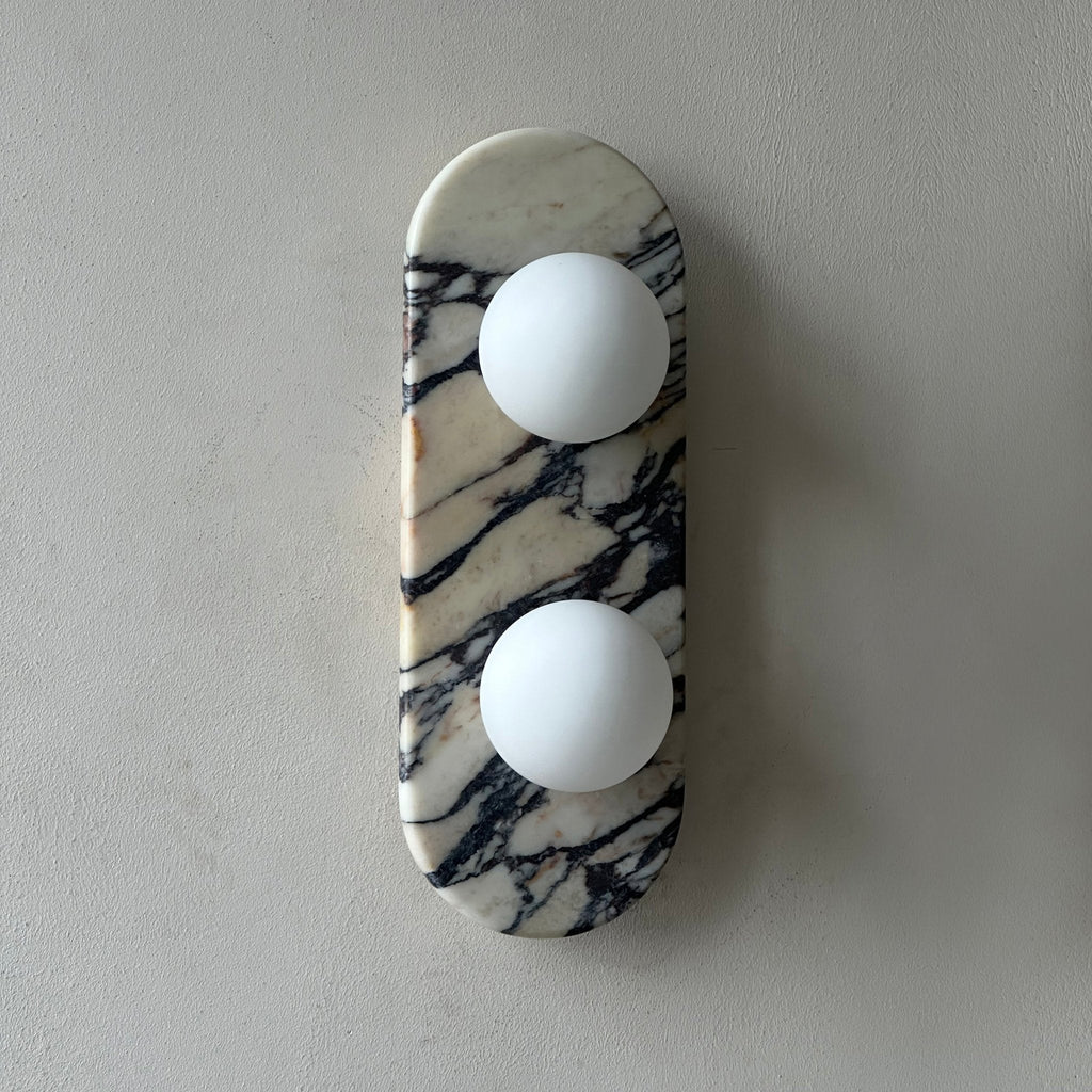 Clérachic marble wall light unlit, photographed close-in on a grey wall with the marble veining sharply visible.
