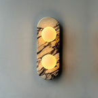 Clérachic marble wall light lit warm on a plain grey wall, photographed in soft focus with a gentle glow around it.