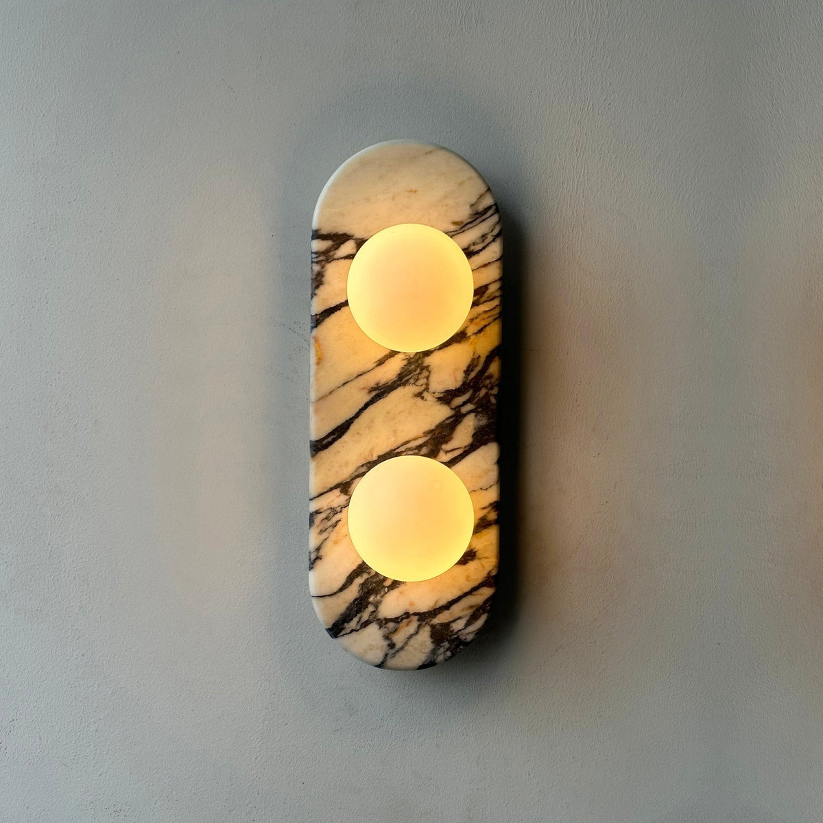 Clérachic marble wall light lit warm on a plain grey wall, photographed in soft focus with a gentle glow around it.
