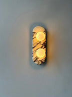 Clérachic marble wall light lit warm on a blue-grey wall, mounted higher in frame with wide plain wall around it.