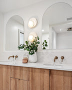 Between two arched mirrors in a wood-vanity bathroom, stacked Clérachic wall lights sit above brass taps and a leafy plant.