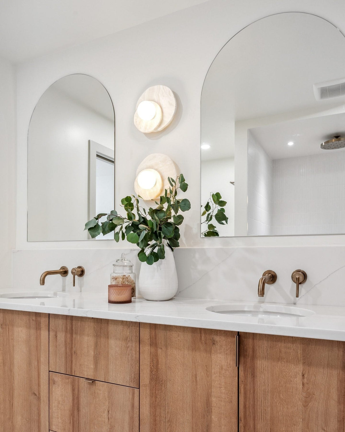 Between two arched mirrors in a wood-vanity bathroom, stacked Clérachic wall lights sit above brass taps and a leafy plant.