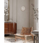 On a panelled wall beside dried branches, a Clérachic wall light hangs near a wood dresser and a leather-strap lounge chair.