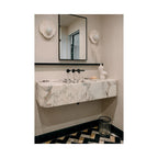 Two Clérachic wall lights flank a mirror above a ribbed marble sink in a powder room with dark patterned floor tiles.