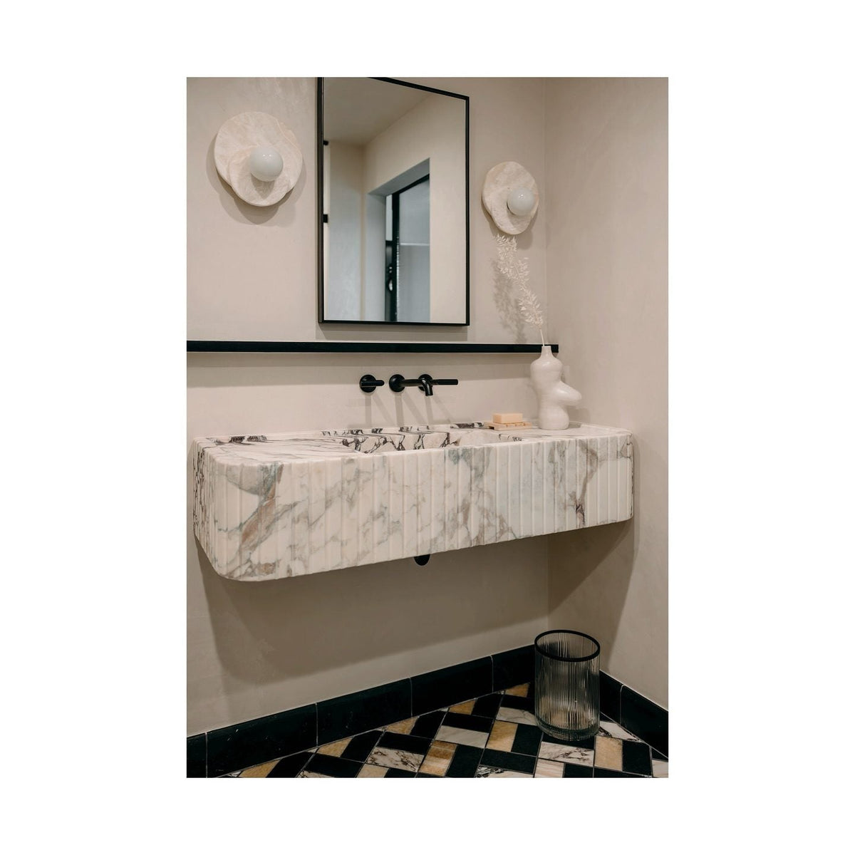 Two Clérachic wall lights flank a mirror above a ribbed marble sink in a powder room with dark patterned floor tiles.