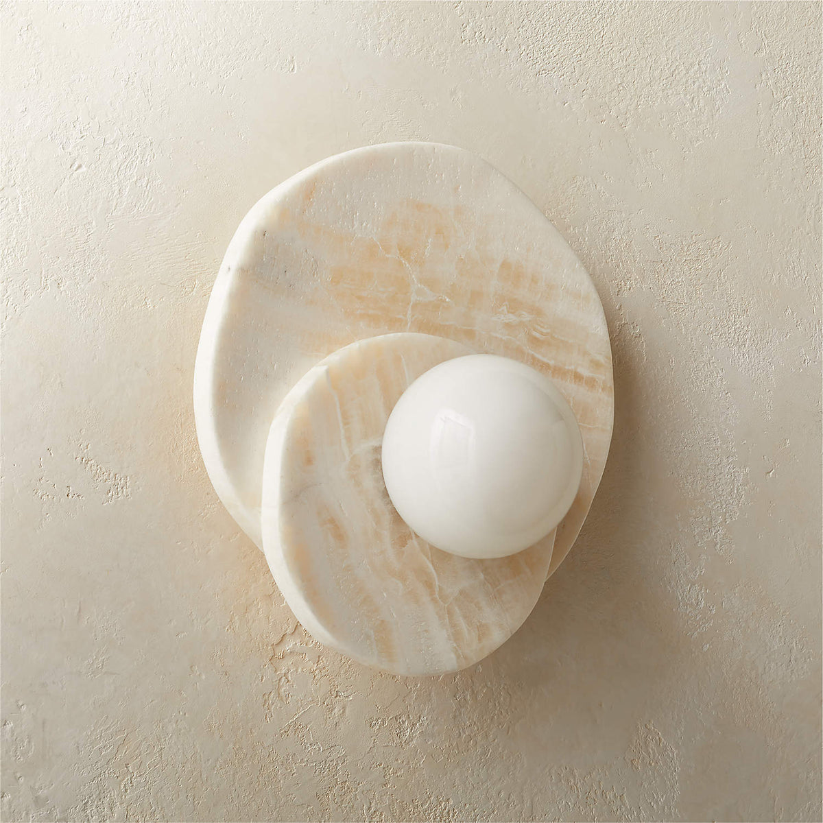 Front-on against a rustic beige wall, the alabaster Clérachic wall light's stacked discs catch warm natural daylight.