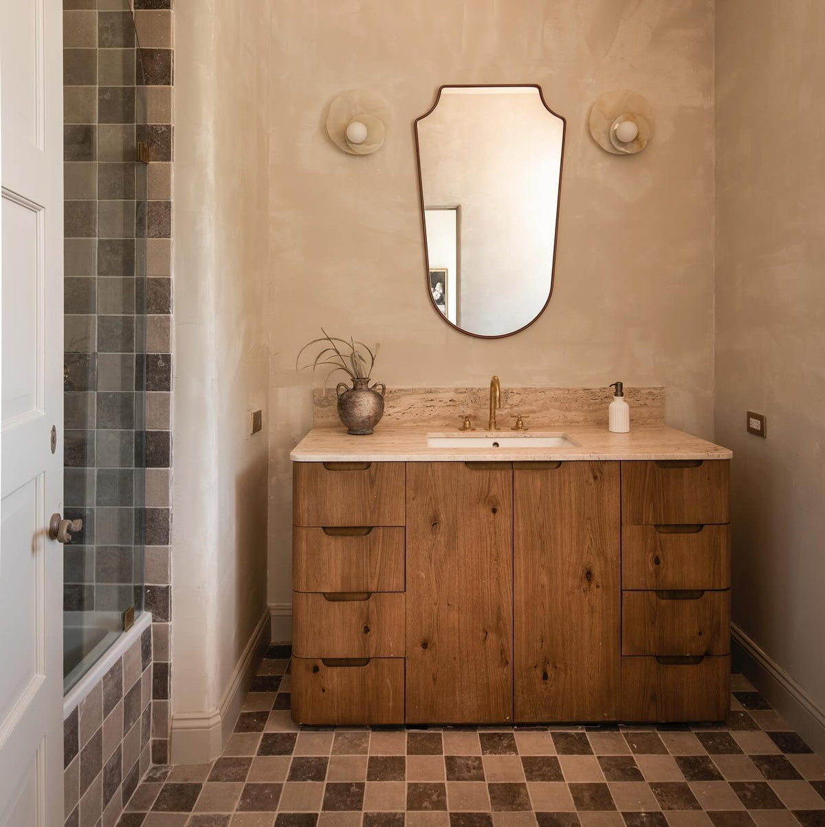 Flanking a bathroom mirror above a wood vanity, two Clérachic wall lights sit on a plaster wall over checked tile flooring.